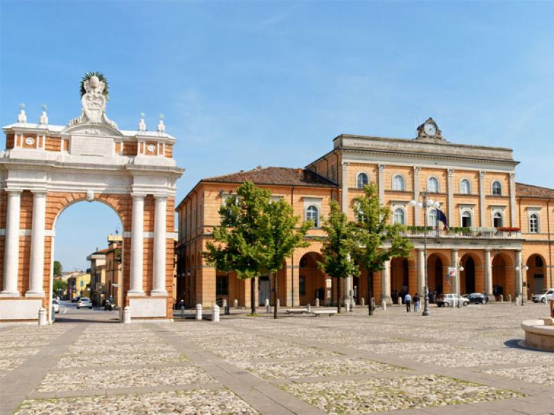 Piazza con fontana, arco storico e edificio in mattoni, cielo sereno.